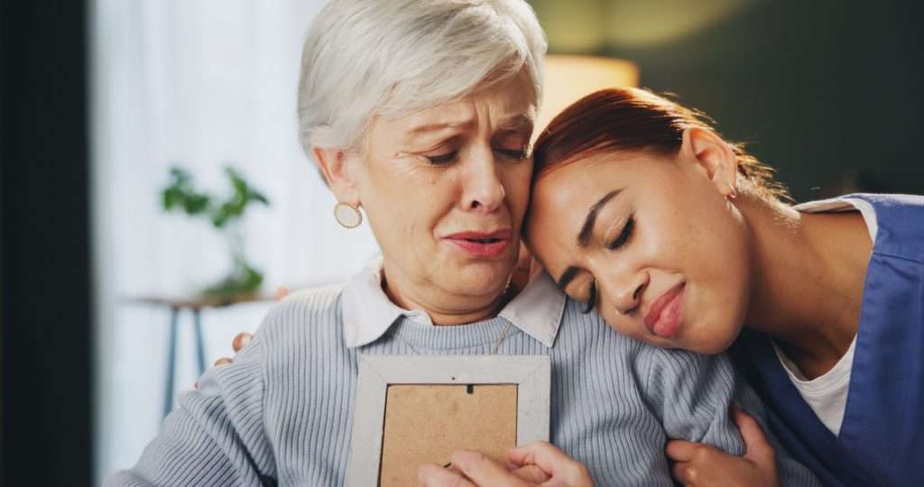 Senior woman, sad and grief in retirement home with caregiver, picture frame and reflection as pens